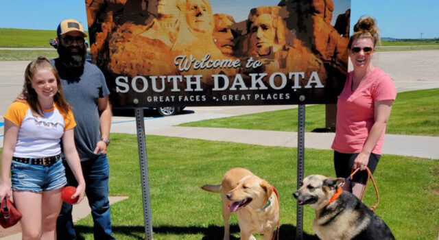 Clark family stands in front of Welcome to South Dakota sign with two dogs