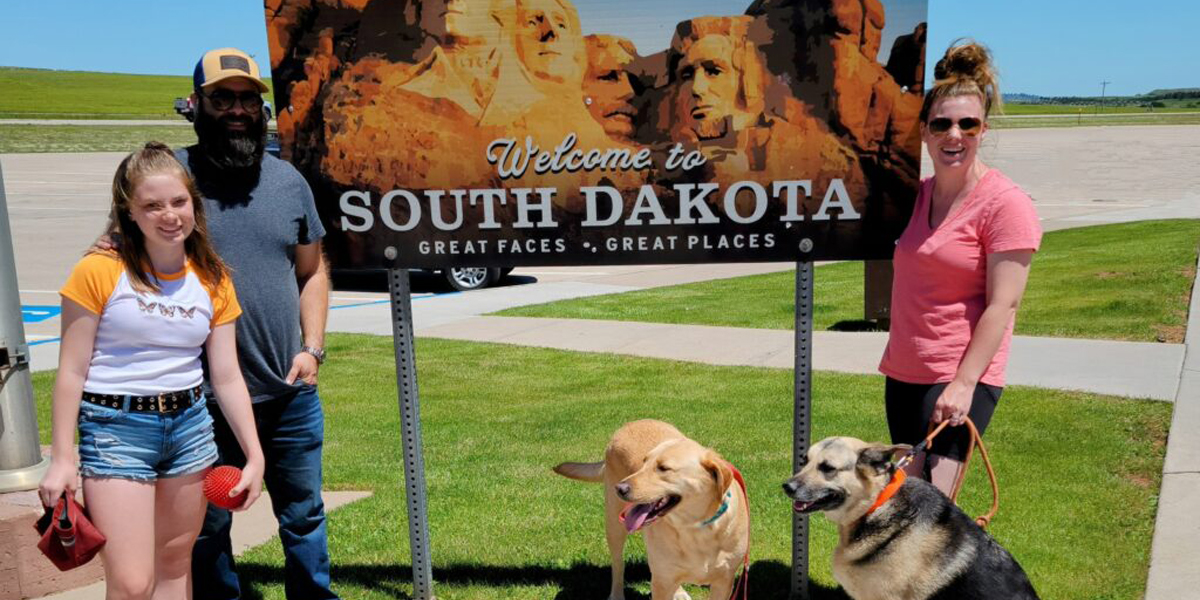 Clark family stands in front of Welcome to South Dakota sign with two dogs