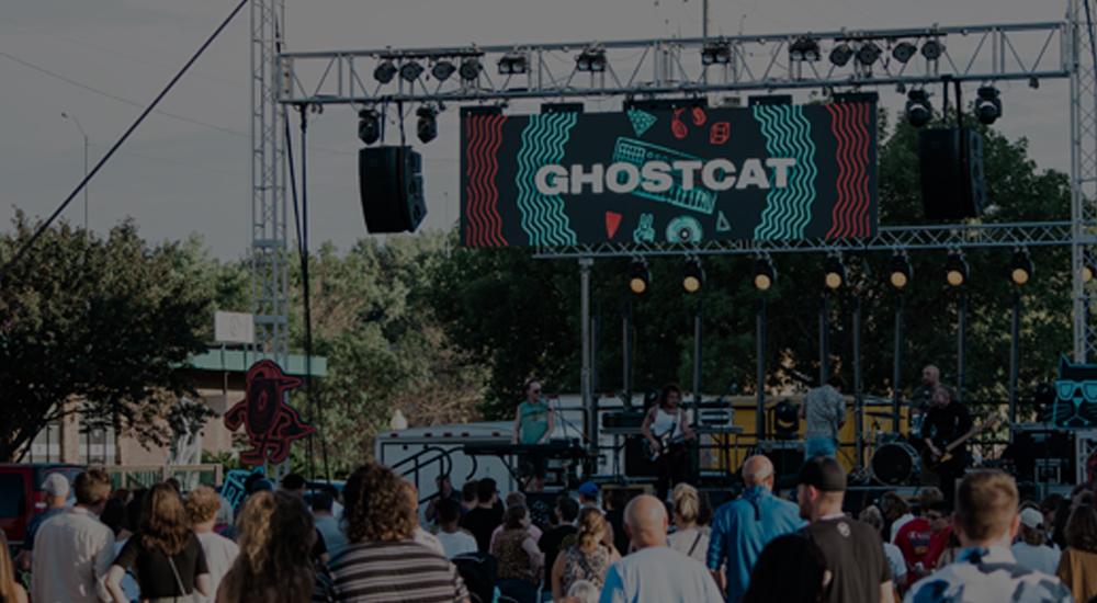 A photo of a band playing in Downtown Sioux Falls, SD, with a sign that says 'Ghostcat' above them